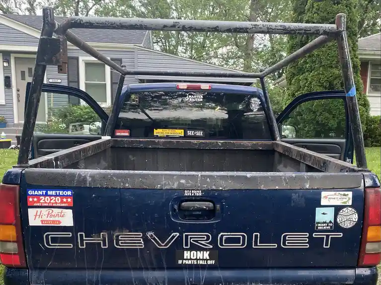 The rear view of a blue Chevrolet pickup truck covered in various stickers, including one that reads 'HONK IF PARTS FALL…