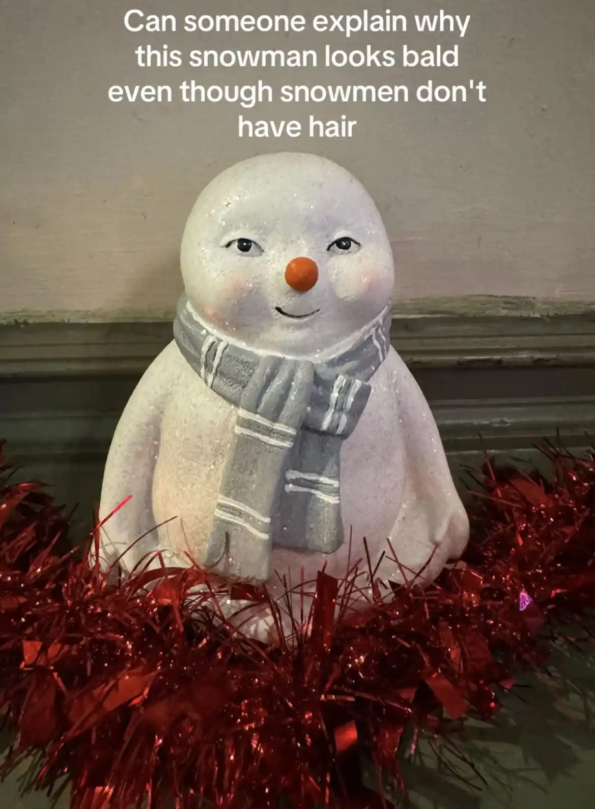 A decorative snowman figurine with a round head, orange carrot nose, and gray striped scarf sits atop red tinsel. The…