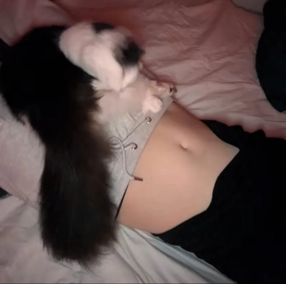A person lying on their back on a bed with pink sheets, wearing a grey hoodie with the drawstring undone, exposing their…