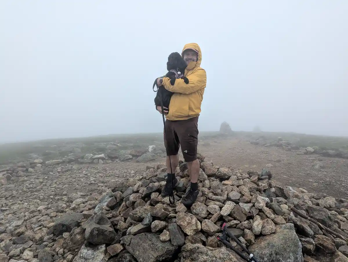 A man in a yellow rain jacket stands atop a rocky mountain summit, holding a small black dog in his arms. The scene is…