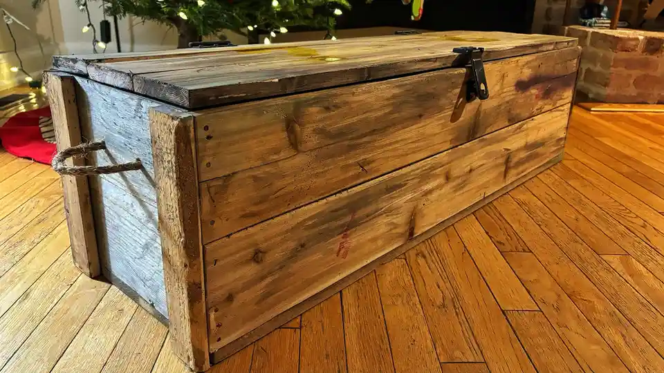 A rustic, weathered wooden chest with a rope handle and metal latch sits on a hardwood floor, with a lit Christmas tree…