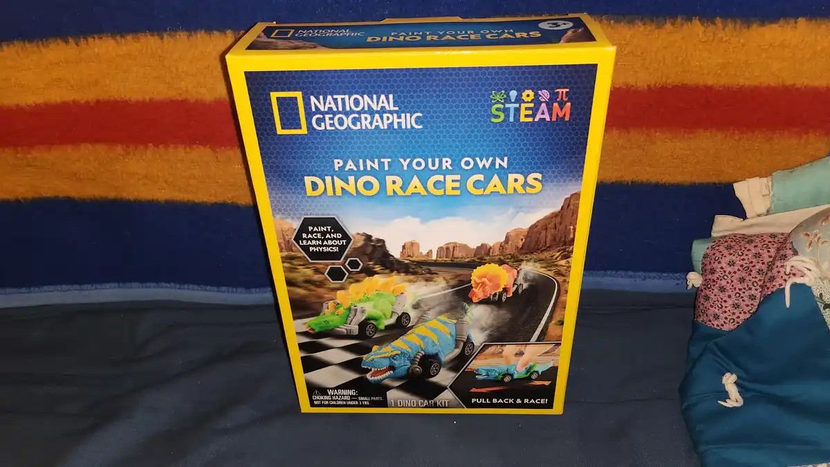 A yellow box for a National Geographic 'Paint Your Own Dino Race Cars' toy kit sits on a striped blanket. The box art shows…
