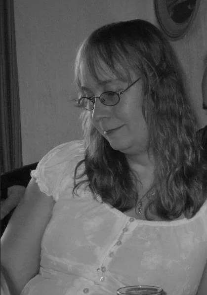 A black-and-white photo of a woman with glasses and long hair, wearing a white blouse, looking down with a slight smile…