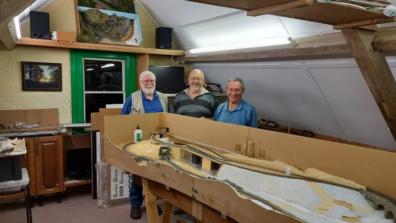 three smiling older men stand behind a large, unfinished model train layout built on a wooden frame in a cluttered workshop…