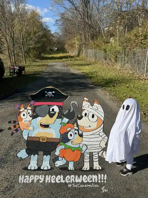 A child in a white ghost costume stands beside a large chalk drawing on a paved path of the Bluey characters dressed in…