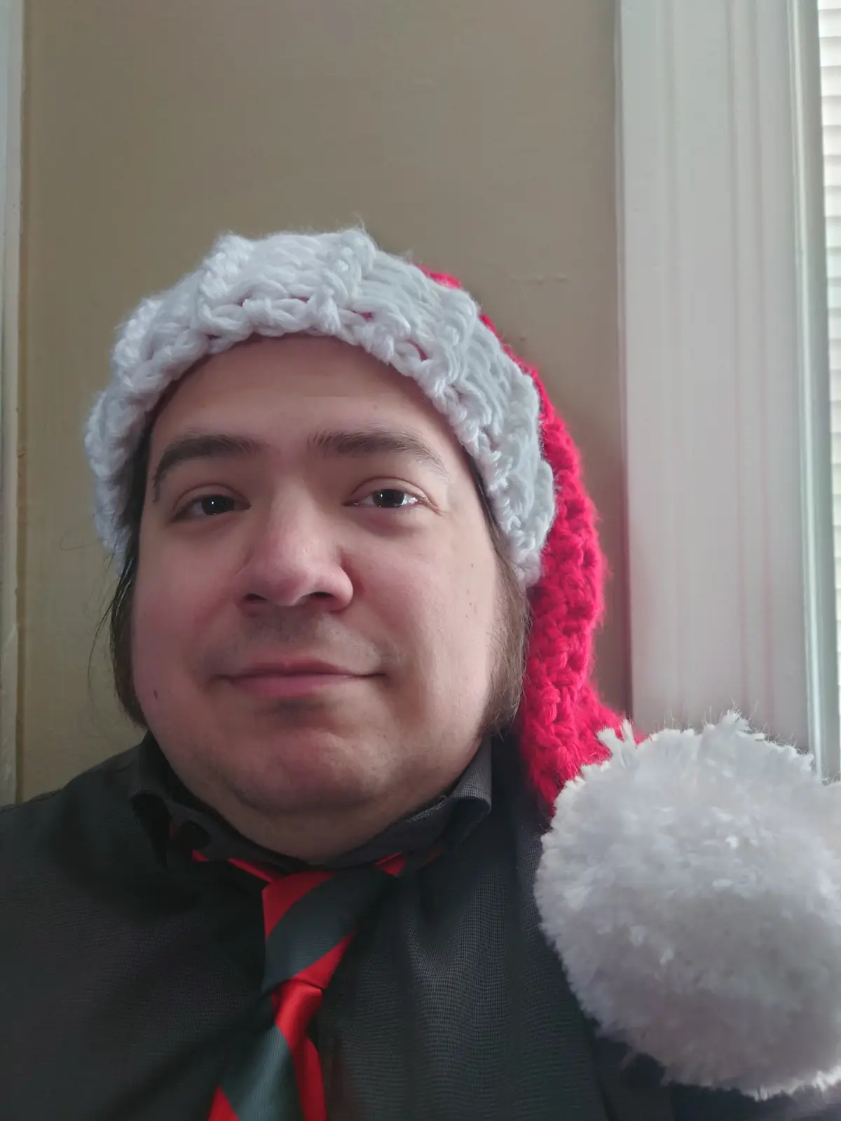 A man with long brown hair wears a red and white crocheted Santa hat with a fluffy white pom-pom, paired with a dark…
