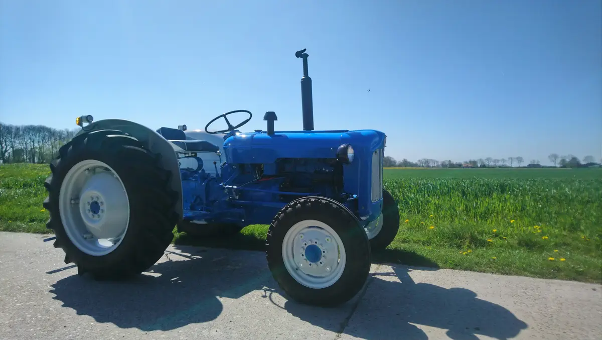A bright blue vintage tractor with large black tires and white rims is parked on a paved road beside a green field under a…