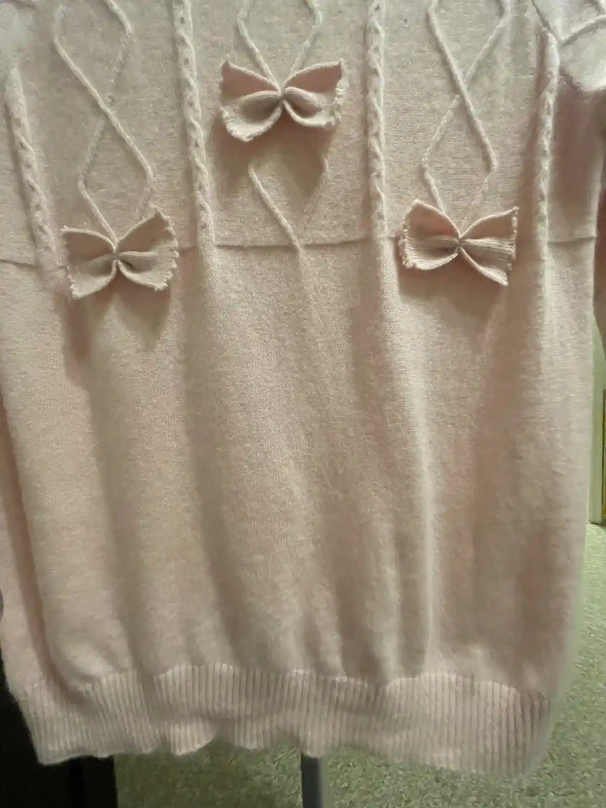 A close-up photo shows a light pink knitted sweater with three small, knitted bows sewn onto the front, positioned…
