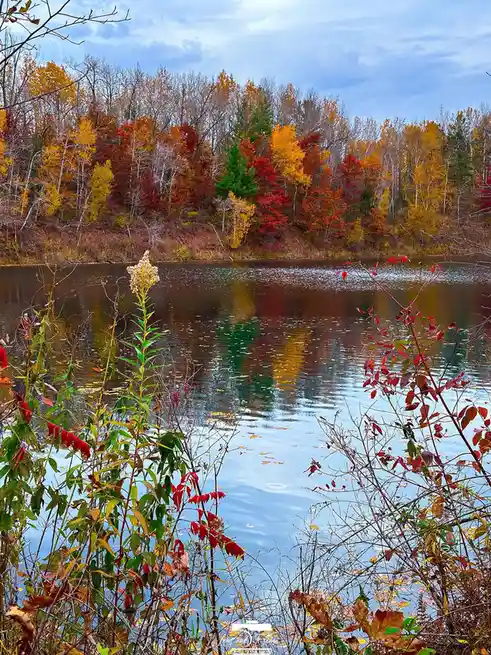 A serene autumn landscape featuring a calm lake reflecting a forest of vibrant orange, red, and yellow trees under a partly…