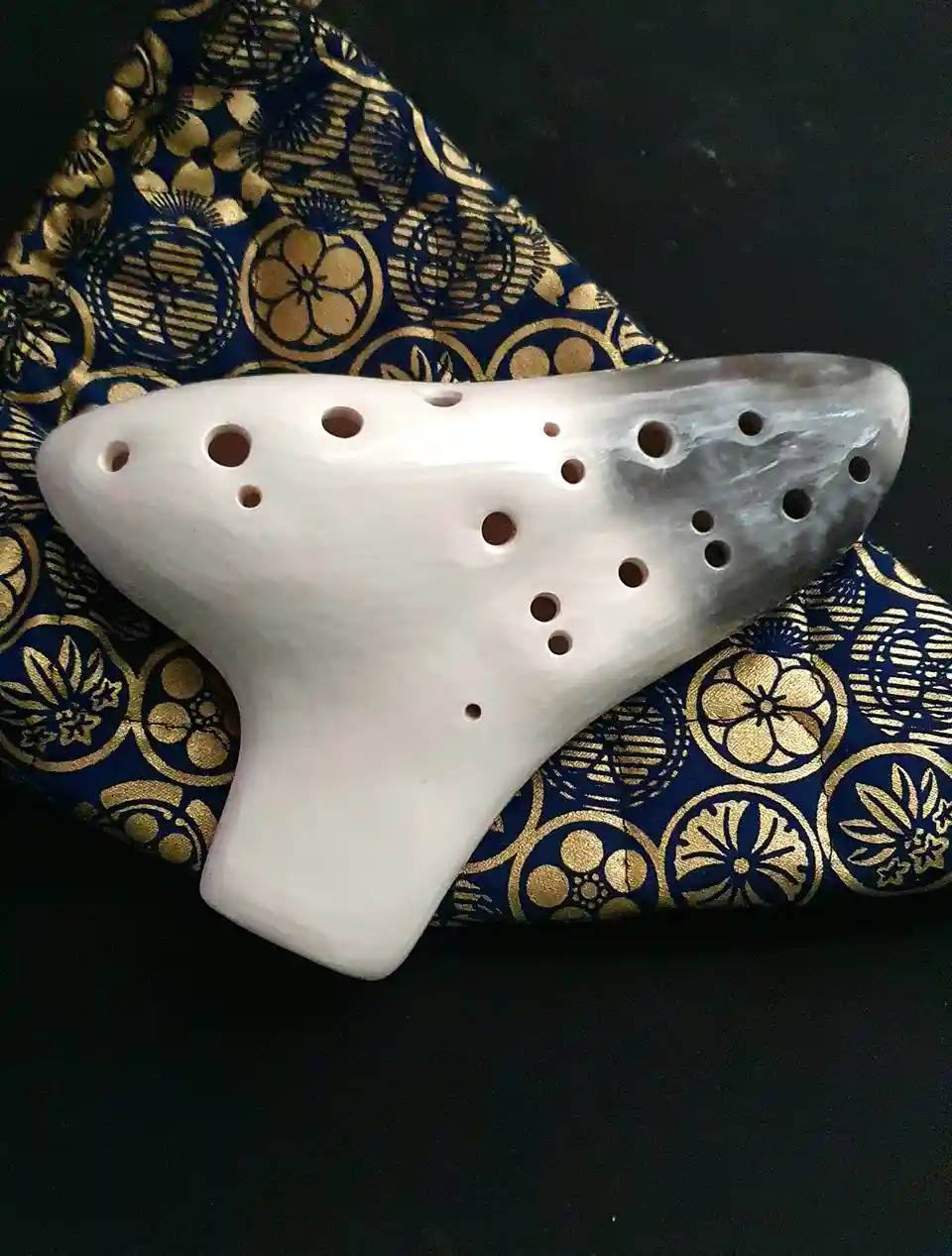 A white ceramic ocarina with multiple finger holes rests on a dark blue fabric with gold floral patterns. The ocarina's…