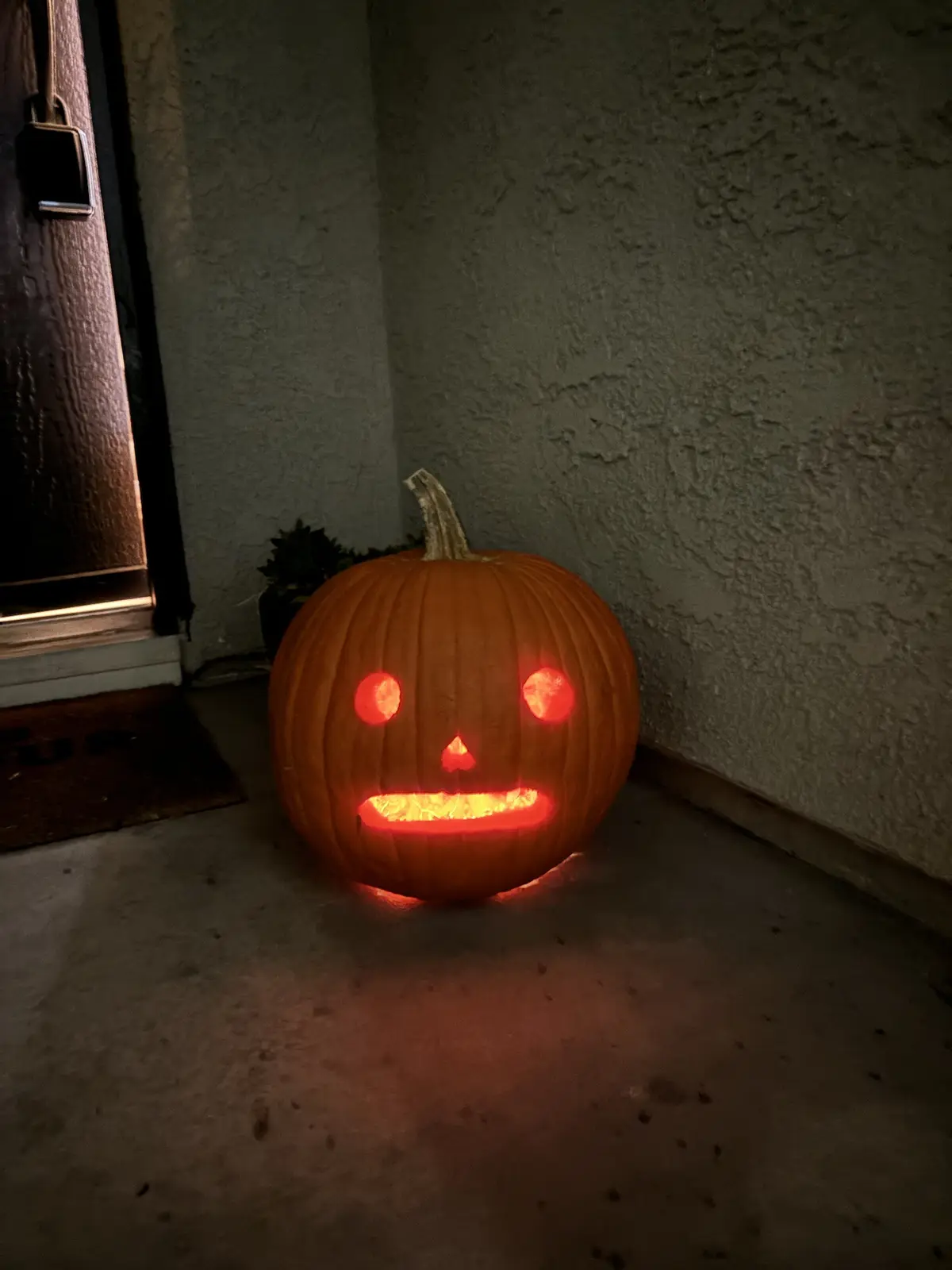 A carved orange pumpkin with glowing red eyes, nose, and mouth sits on a concrete porch near a door, illuminated from within…