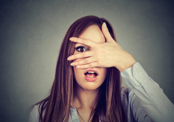 a woman with long brown hair, wearing a light-colored button-up shirt, covers her eyes with her right hand while her left…