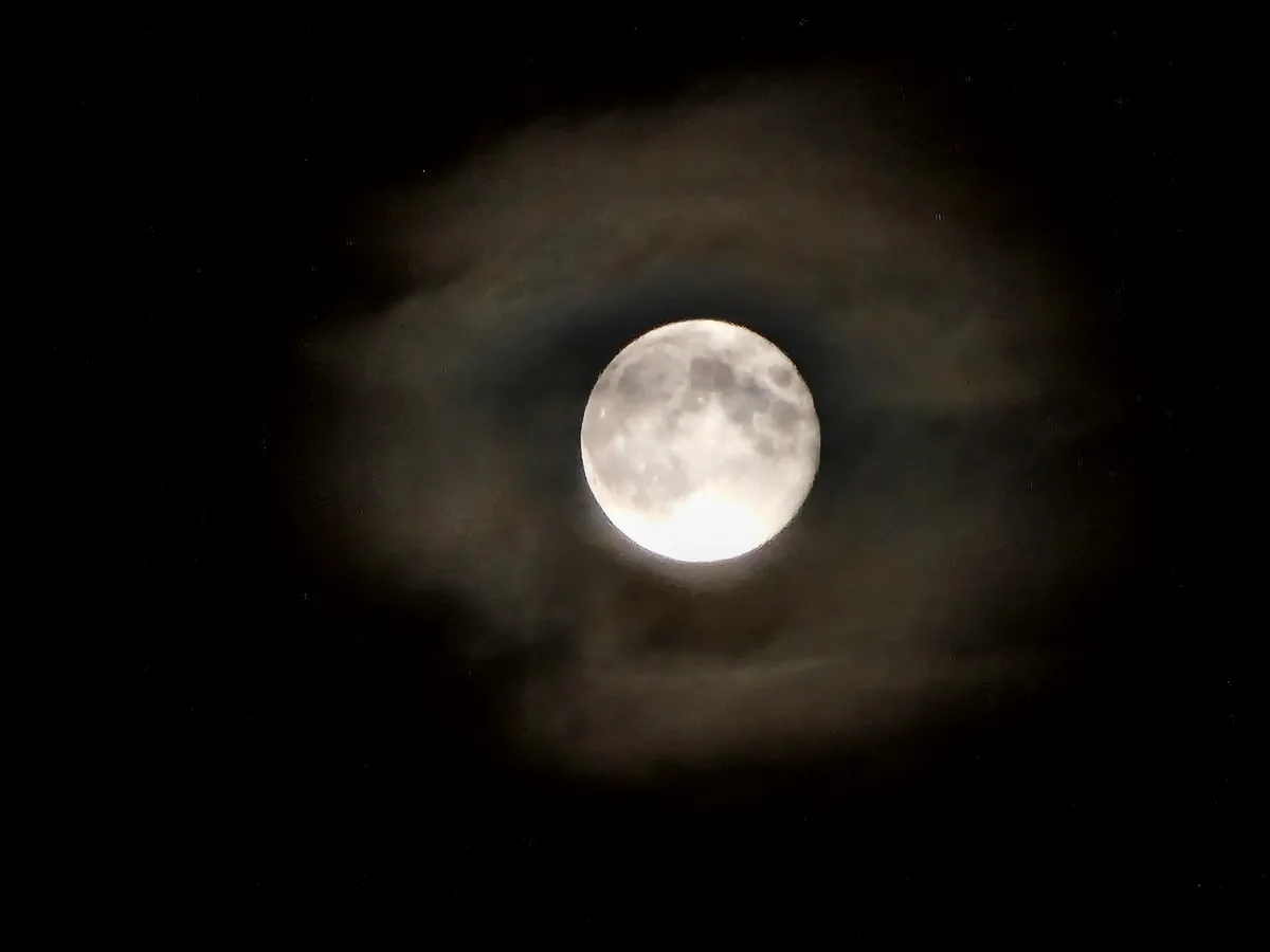 A full moon is visible through a dark, cloudy sky, with the moon's light illuminating the surrounding clouds. The overlaid…