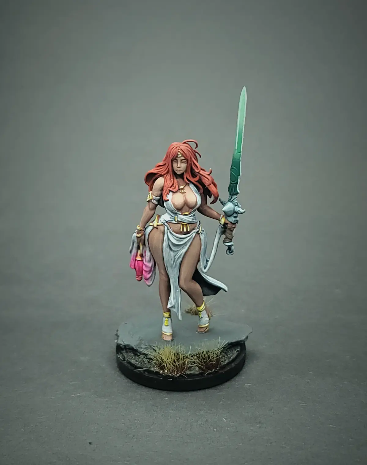 A painted miniature figurine of a female warrior with long red hair, holding a green sword and a pink object, standing on a…