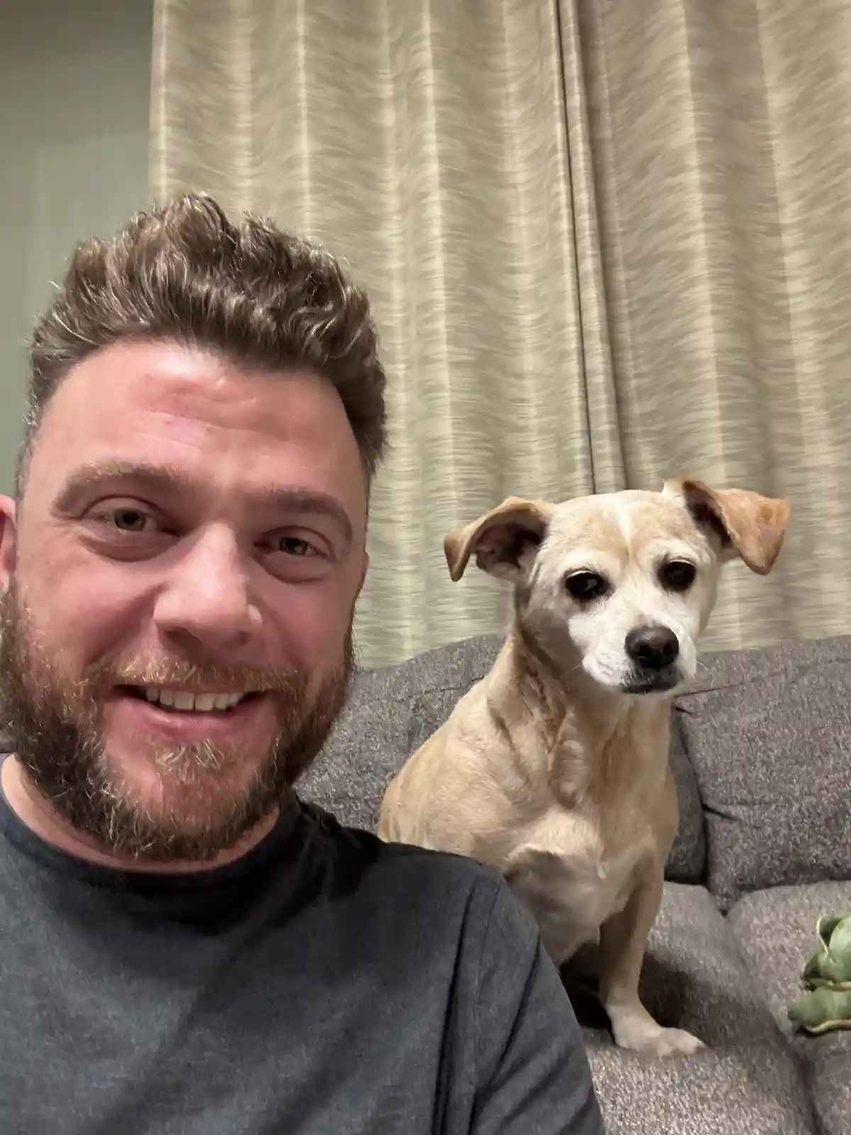 A man with a beard and dark shirt smiles at the camera while sitting on a gray couch next to a light-colored dog with floppy…