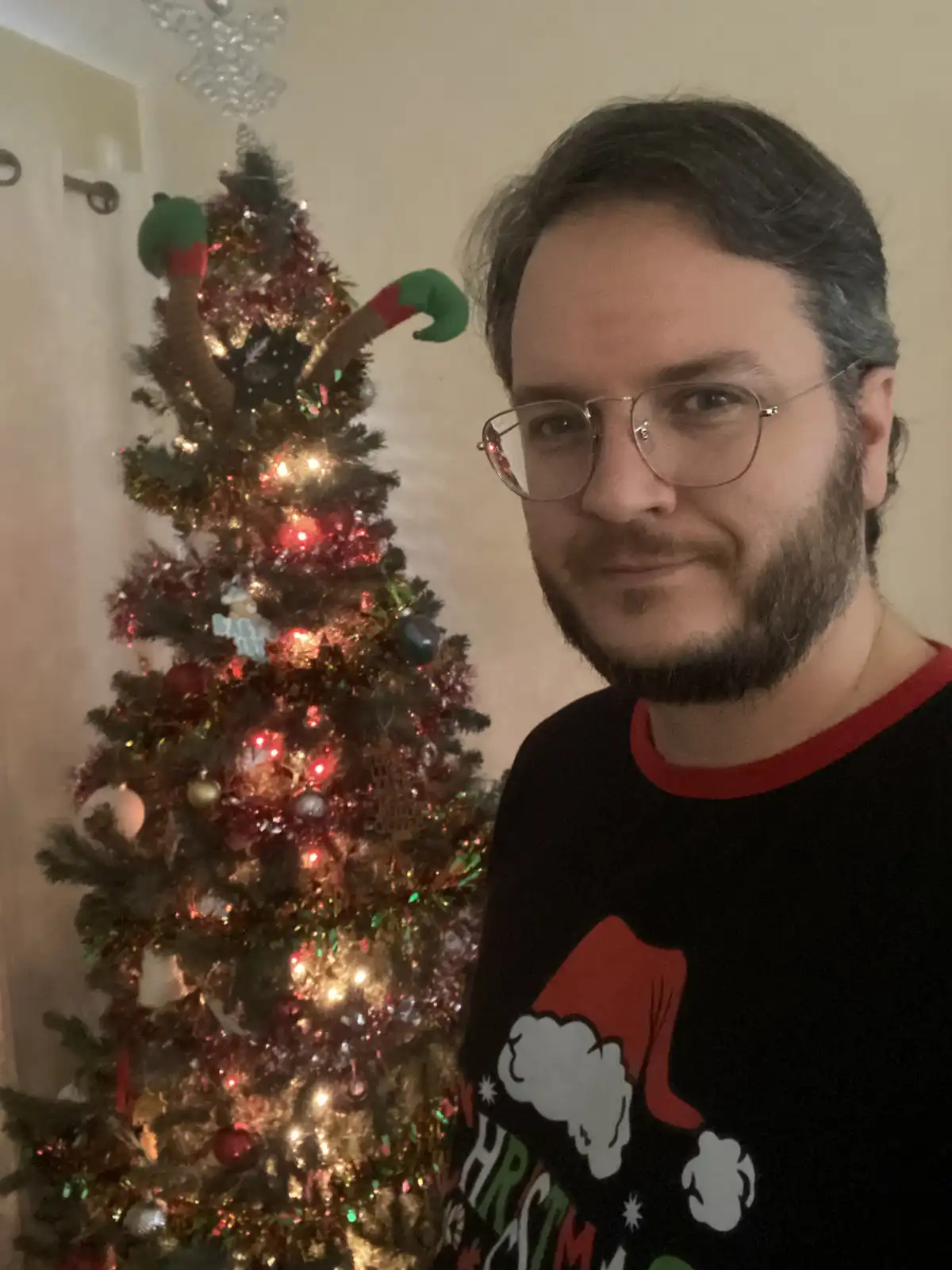 A man with glasses and a beard wearing a black Christmas-themed shirt stands in front of a decorated Christmas tree. The…