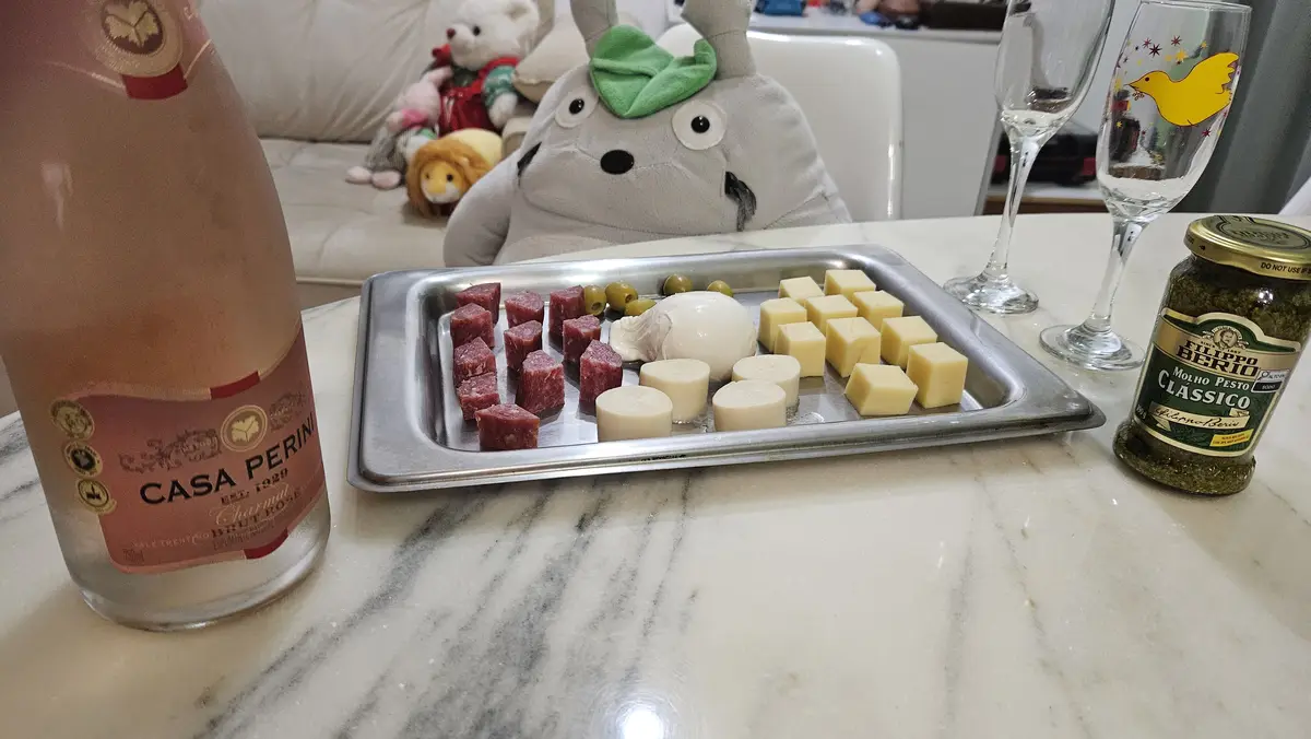 A plush Totoro plushie sits at a table as if dining, facing a tray of cheese, salami, olives, and burrata. Beside it are a…