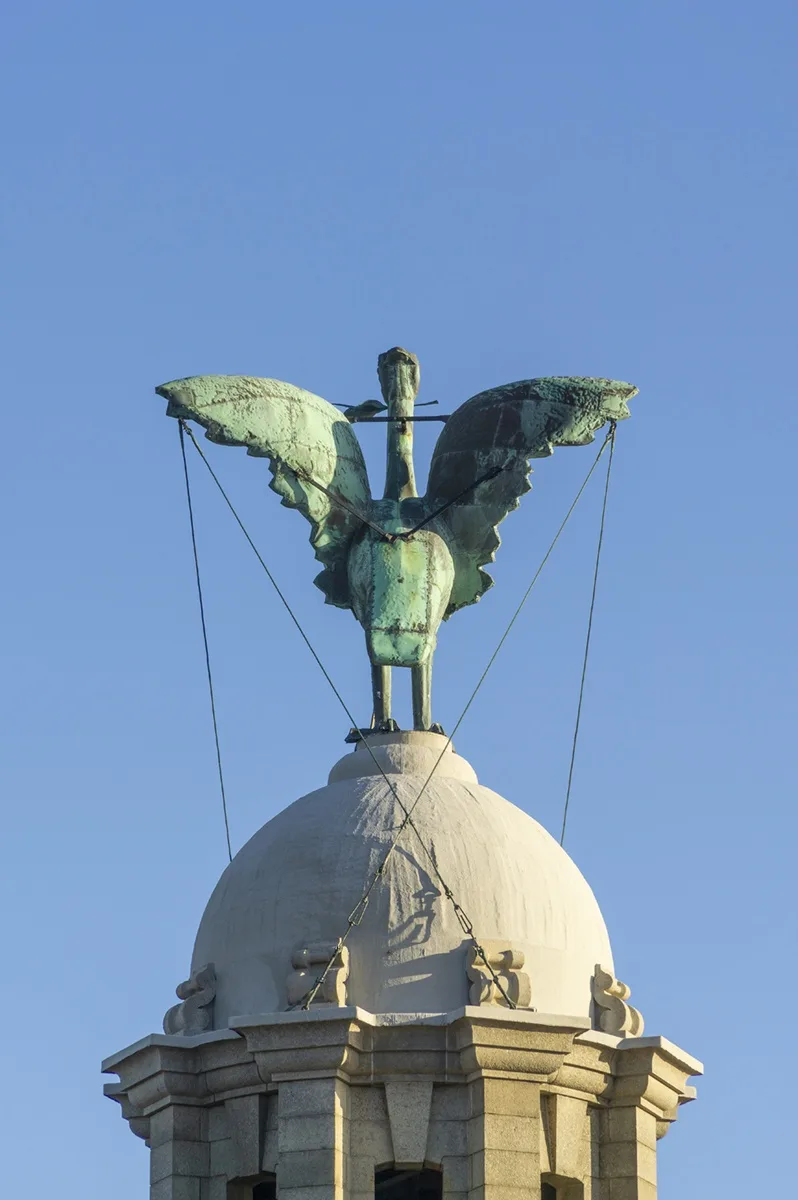 A weathered bronze statue of a bird with outstretched wings perched atop a stone dome, secured by ropes against the clear…
