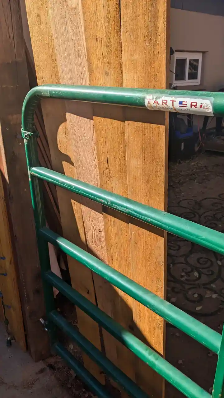 A green metal cattle guard or gate frame leans against a stack of wooden planks outdoors. The top horizontal bar has a faded…