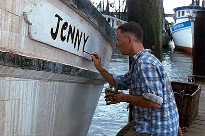 A man in a blue plaid shirt is painting the name 'JENNY' in bold black letters onto the side of a white boat at a dock. He…