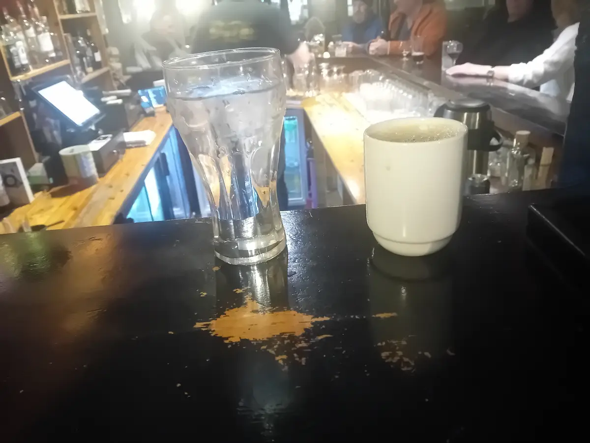 A close-up photo shows a tall, empty beer glass and a white coffee mug sitting side-by-side on a black bar counter with…