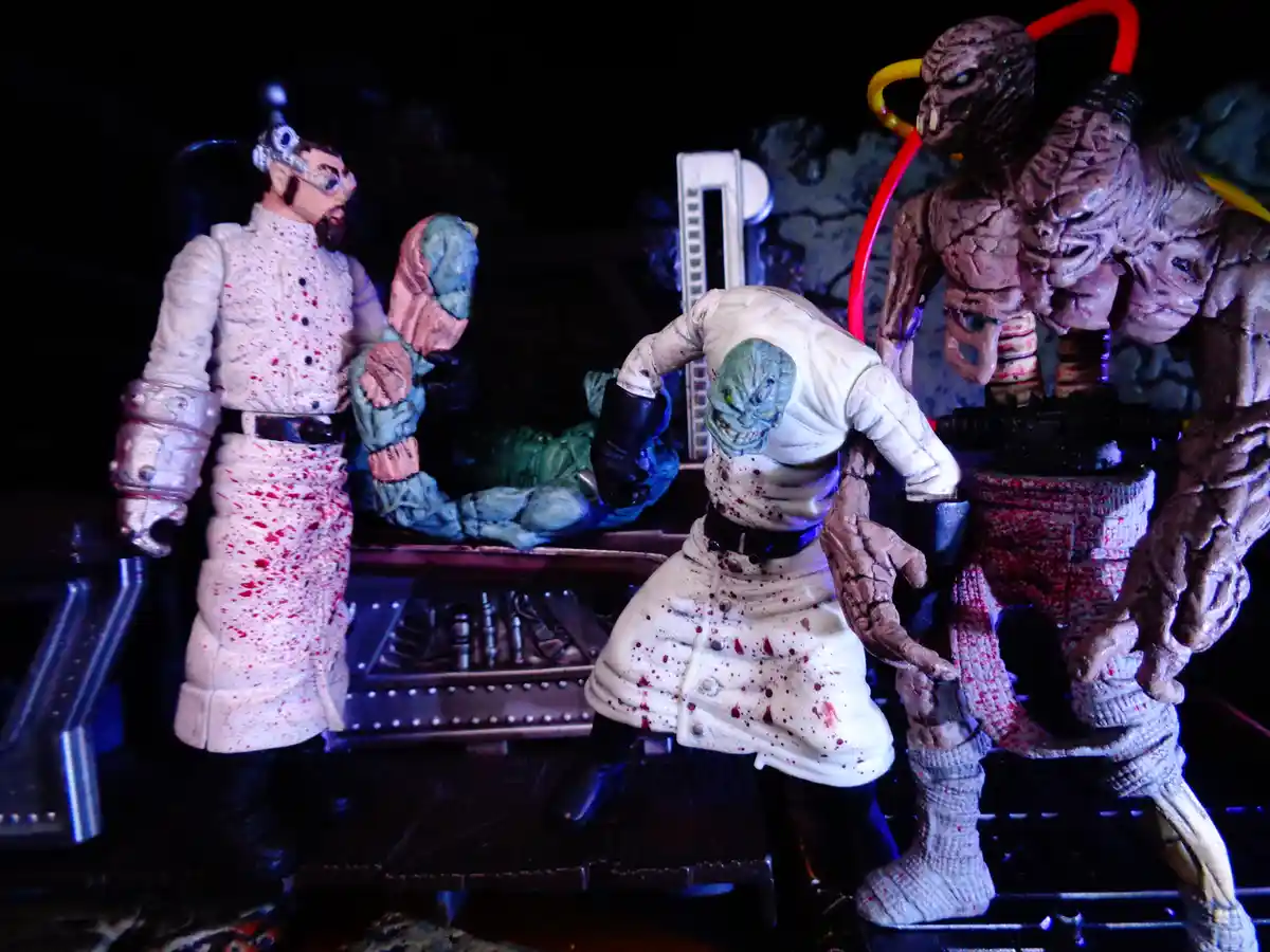 A diorama of three grotesque, blood-splattered action figures in lab coats, resembling mad scientists or alien doctors…