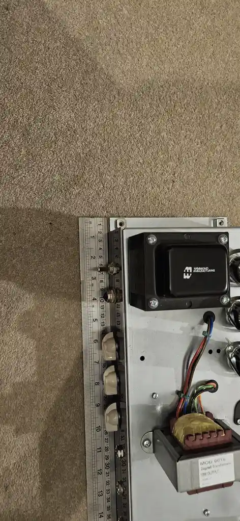 A close-up photo shows the side of an electronic amplifier resting on a beige carpet, with a metal ruler placed vertically…