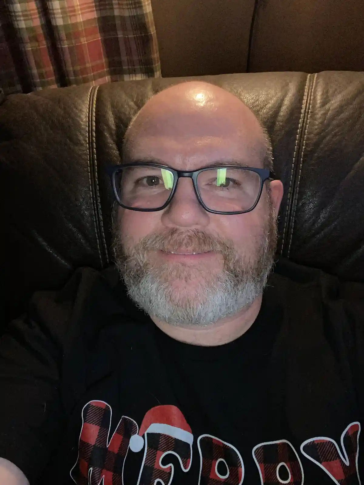 A bald man with a graying beard and glasses smiles while sitting in a dark brown leather recliner, wearing a black t-shirt…