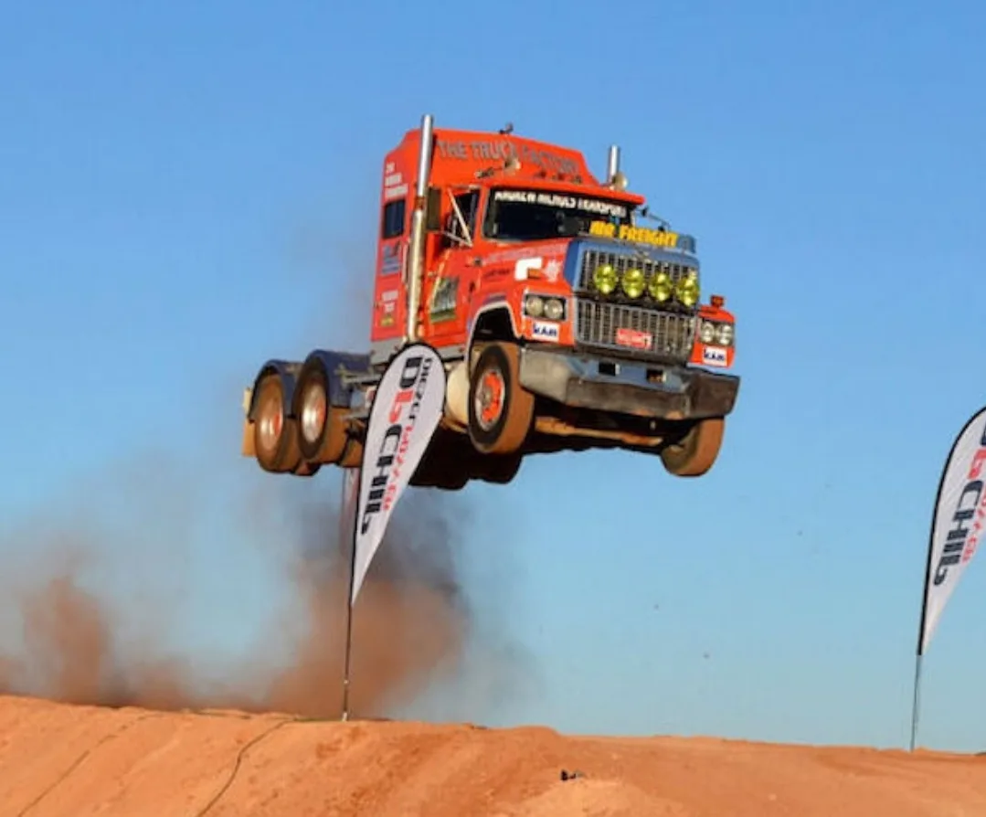 A large orange and blue race truck is airborne over a dirt track, kicking up dust, with two white flags bearing 'B-CHILL'…