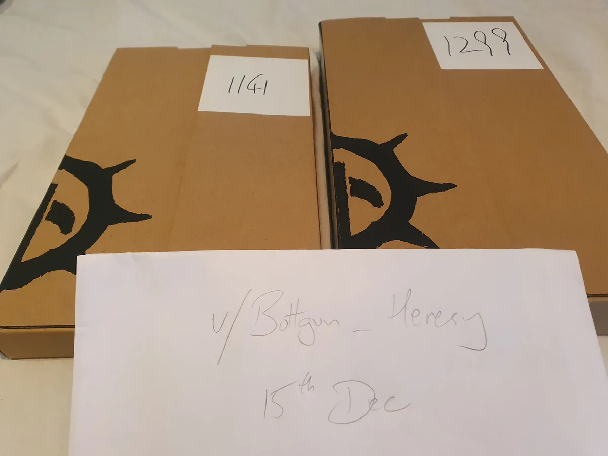 Two brown cardboard boxes with black stylized 'D' logos and handwritten numbers 1141 and 1299 on white labels. A white paper…