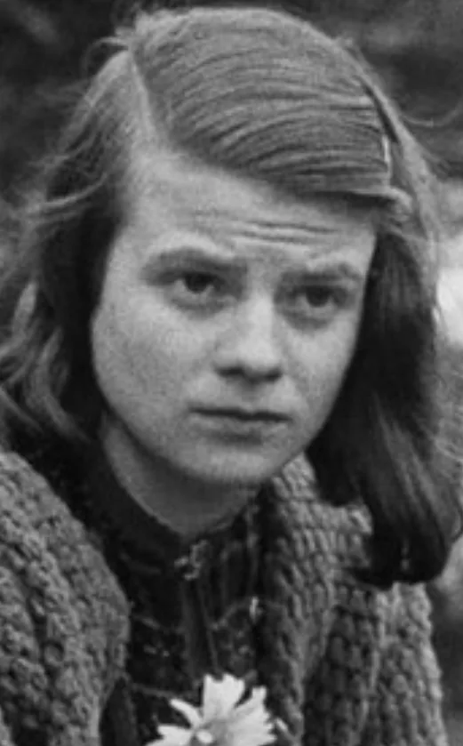 A black-and-white photo of a young person with shoulder-length hair and a knit sweater, looking directly at the camera with…