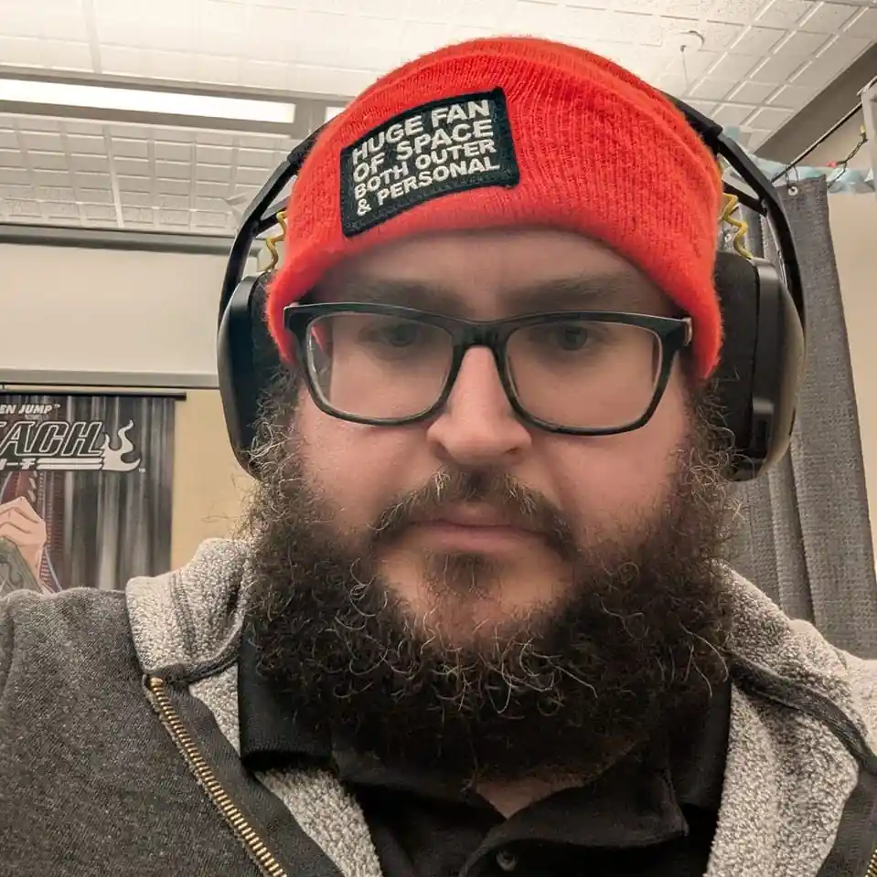 A man with a full beard and glasses wears a bright red beanie with a black patch that reads 'HUGE FAN OF SPACE BOTH OUTER &…