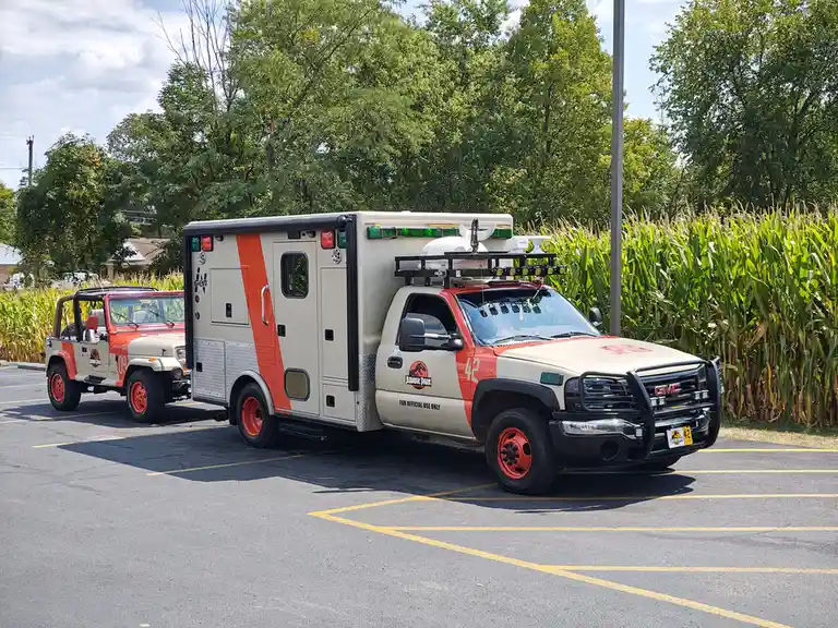 Two vehicles themed after Jurassic Park are parked in a lot: a red and beige GMC truck with a large cargo box and 'FOR…