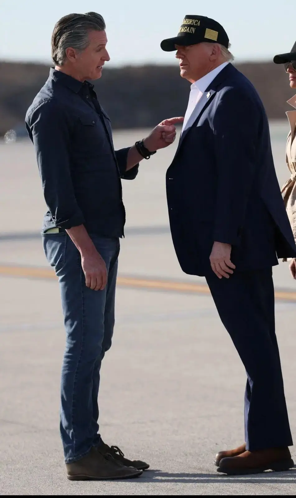 A man in a dark shirt and jeans points at another man in a suit and 'America Again' cap, who stands stoically. The overlaid…