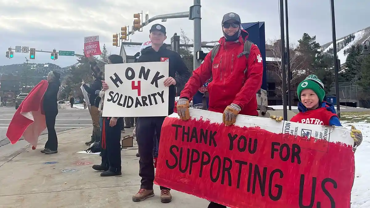 A group of protesters, including a man in a red jacket and a child in a green hat, hold signs on a snowy street. One sign…