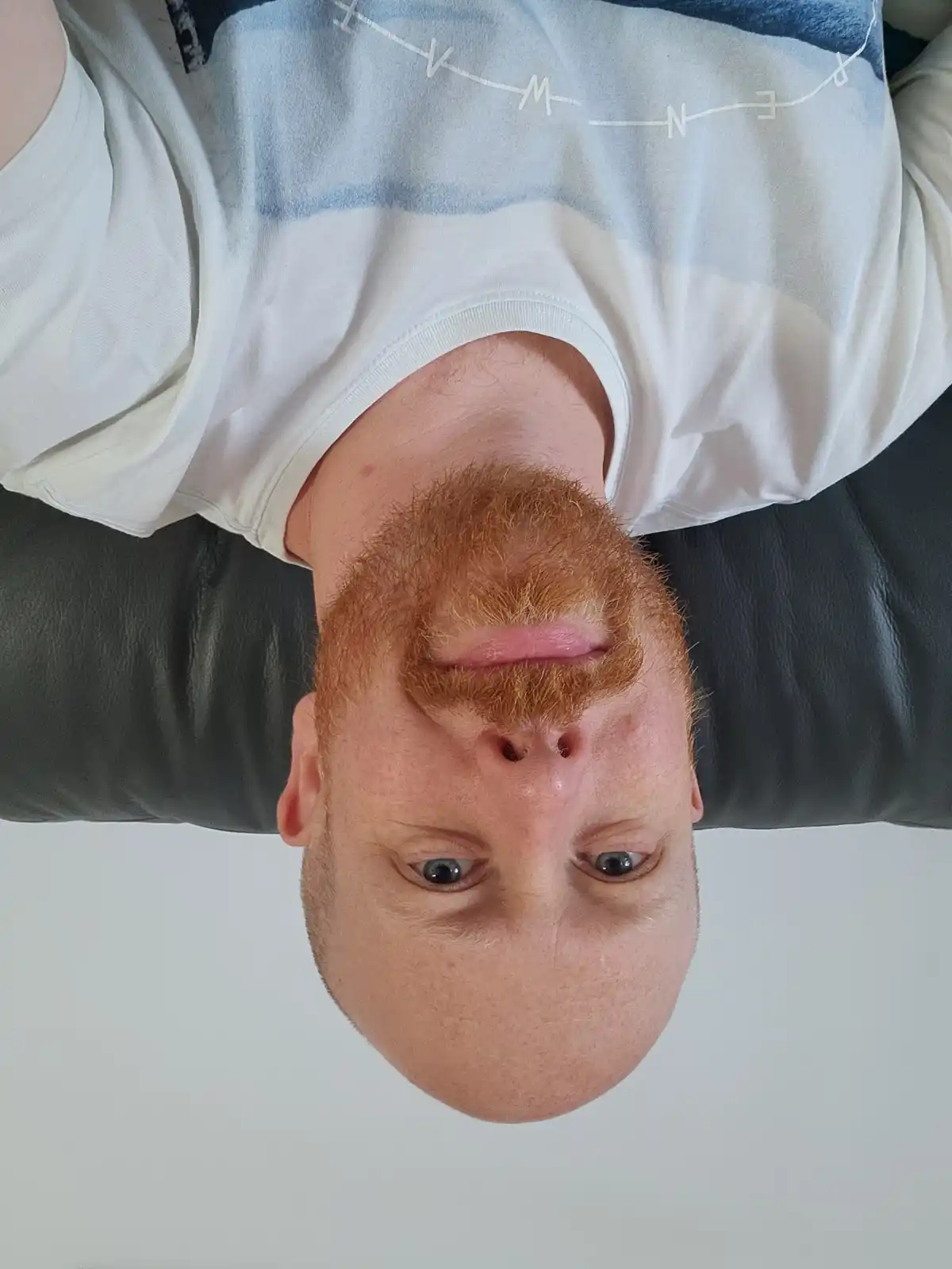 A man with a red beard and blue eyes is photographed upside down, wearing a white t-shirt with a blue and white graphic…