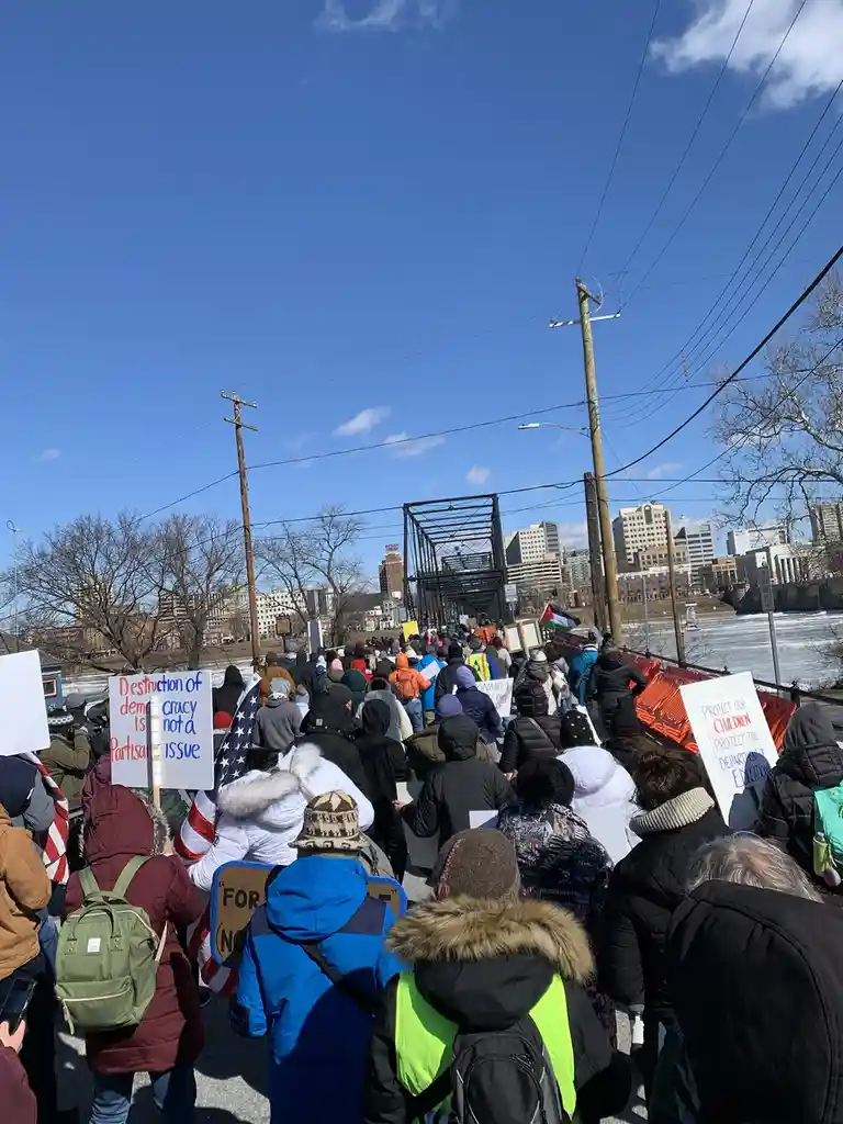 A large crowd of protesters marches across a bridge on a sunny day, many holding signs. One prominent sign reads…