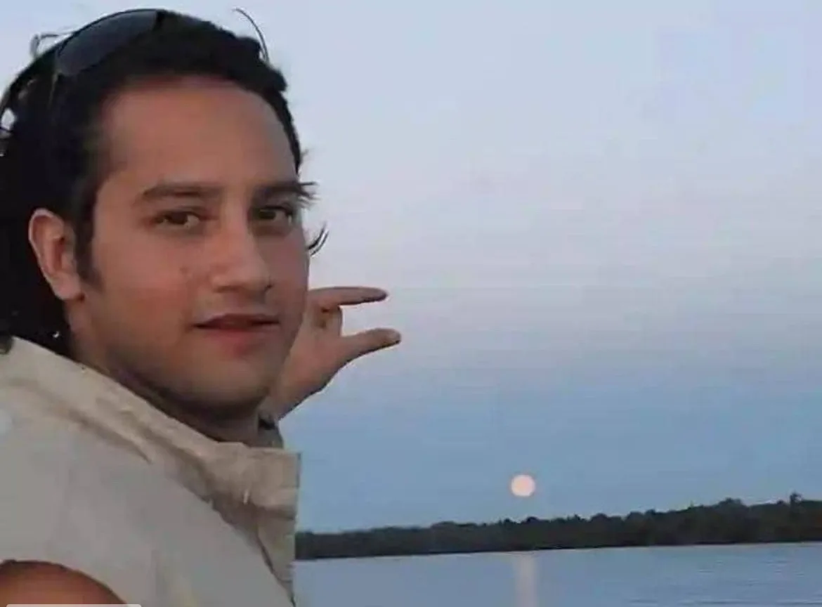 A man with dark hair and sunglasses perched on his head points with his right hand toward a bright, full moon visible over a…