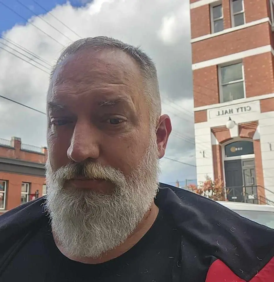 A man with a gray beard and short hair stands in front of a brick building with a sign that reads 'CITY HALL'. The image is…