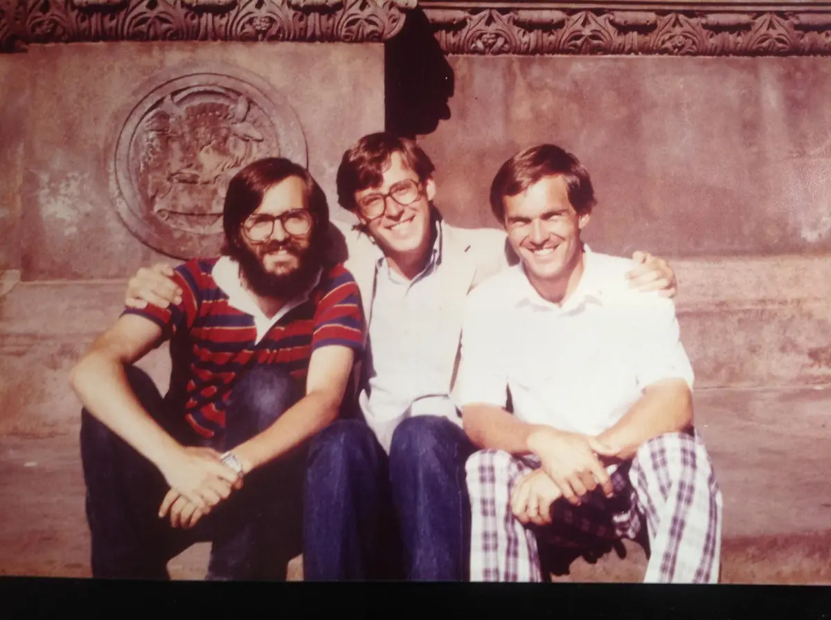 Three men in 1970s-style clothing pose together on a stone bench against an ornate wall with a circular relief. The man on…
