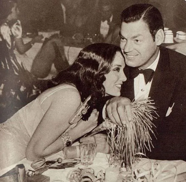 A vintage black-and-white photo shows a man in a tuxedo and a woman in a dress seated at a table, smiling at each other. The…