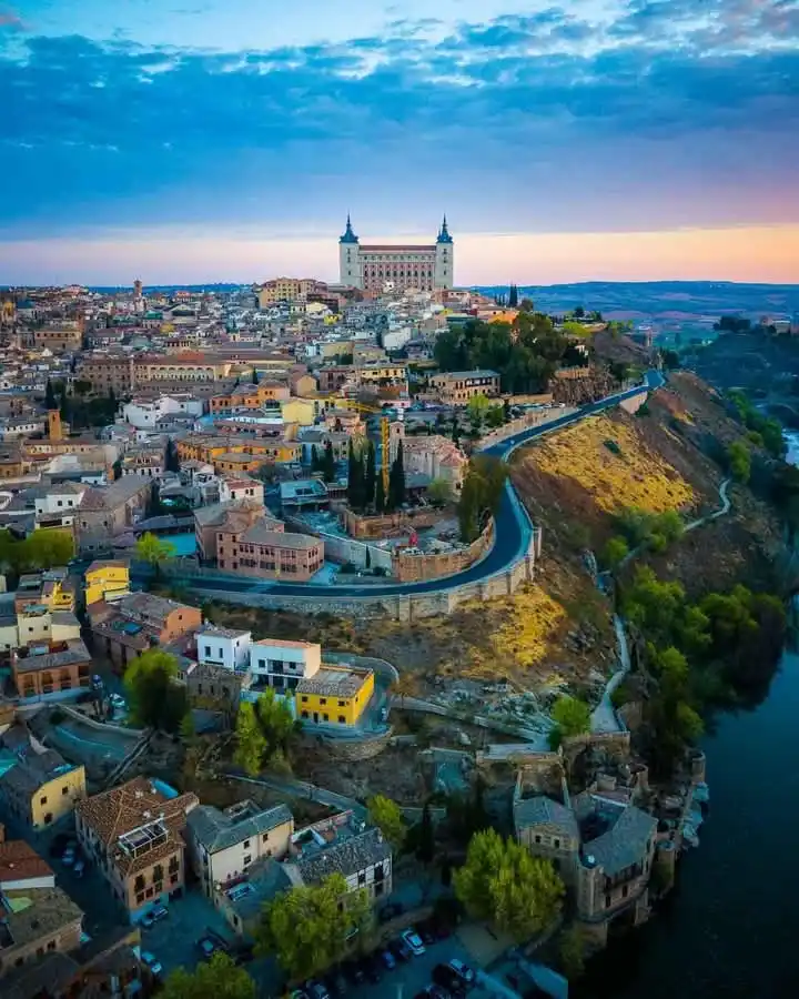 A scenic aerial view of a historic European town at dusk, featuring a prominent castle on a hill overlooking a winding road…
