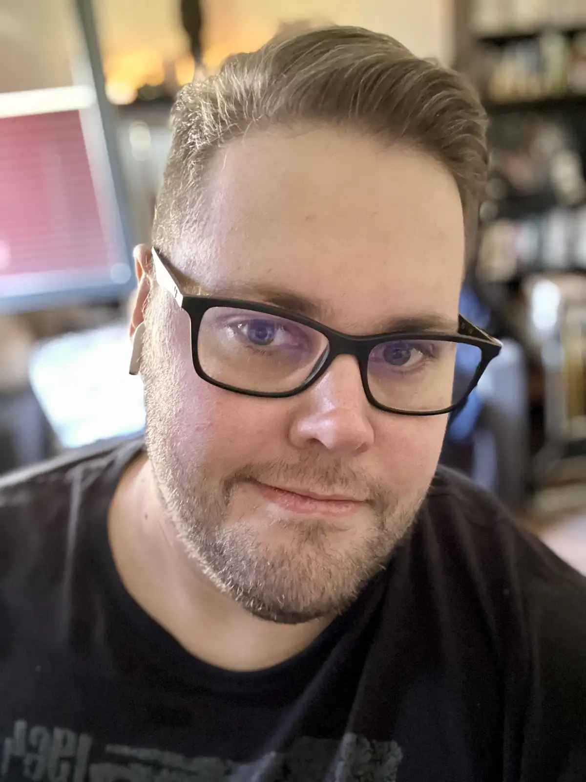A man with glasses and a beard looks directly at the camera with a slight smirk while wearing a white earbud. The overlaid…