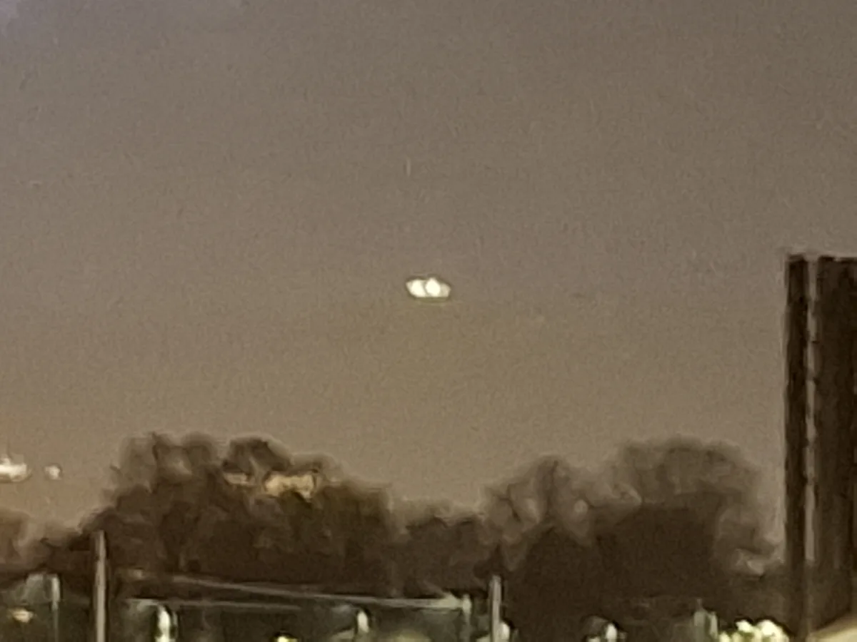 A blurry, sepia-toned nighttime photo shows a faint, glowing, oval-shaped object hovering in the sky above silhouetted trees…