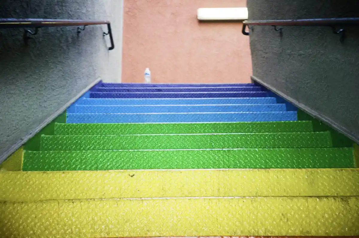 A low-angle shot of a staircase with steps painted in a rainbow gradient from yellow to green to blue to purple, flanked by…
