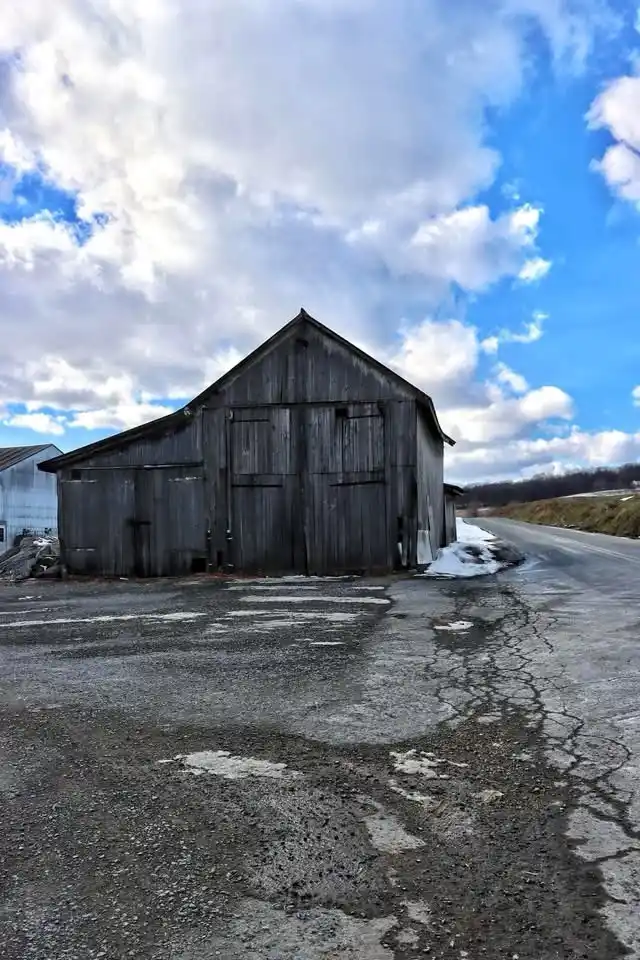 A weathered, gray wooden barn stands alone beside a cracked, wet asphalt road under a partly cloudy blue sky. The ground…