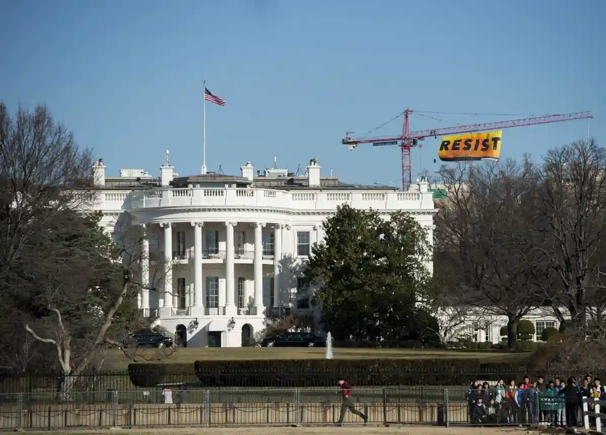 The White House is shown with a red construction crane in the background, holding a large yellow banner with the word…