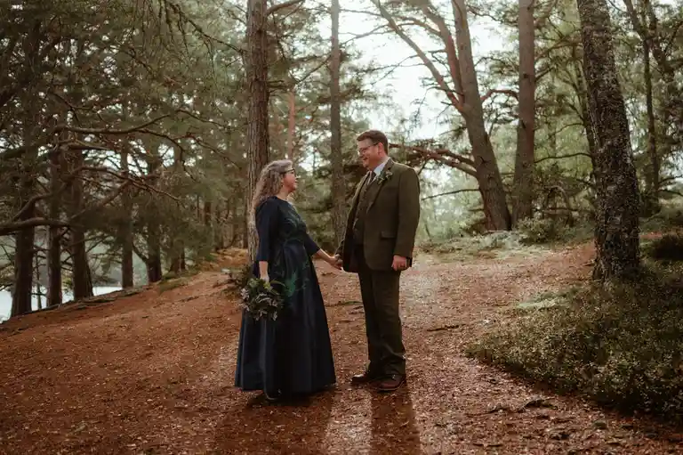 A man and woman in wedding attire stand holding hands on a forest path, surrounded by tall pine trees and fallen needles…