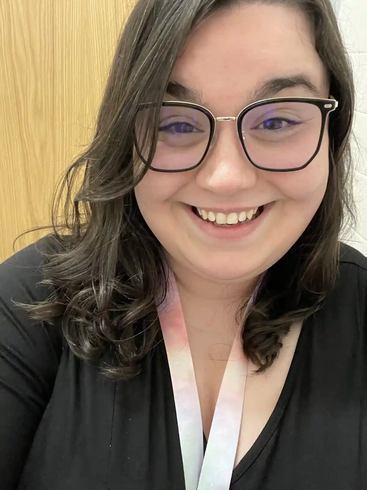 A close-up photo of a smiling woman with dark hair, wearing black-rimmed glasses and a black top, with a colorful lanyard…