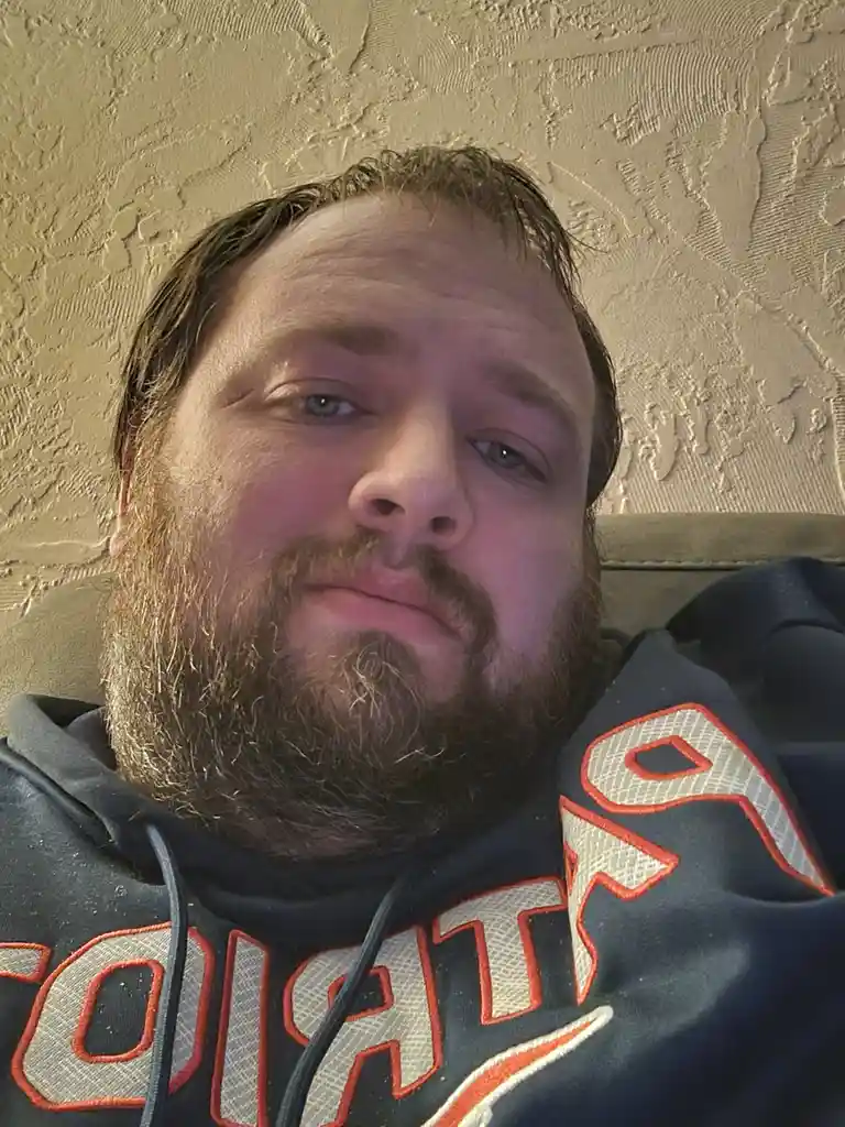 A man with a beard and short hair lies on a couch, wearing a dark hoodie with orange and white lettering. He stares directly…